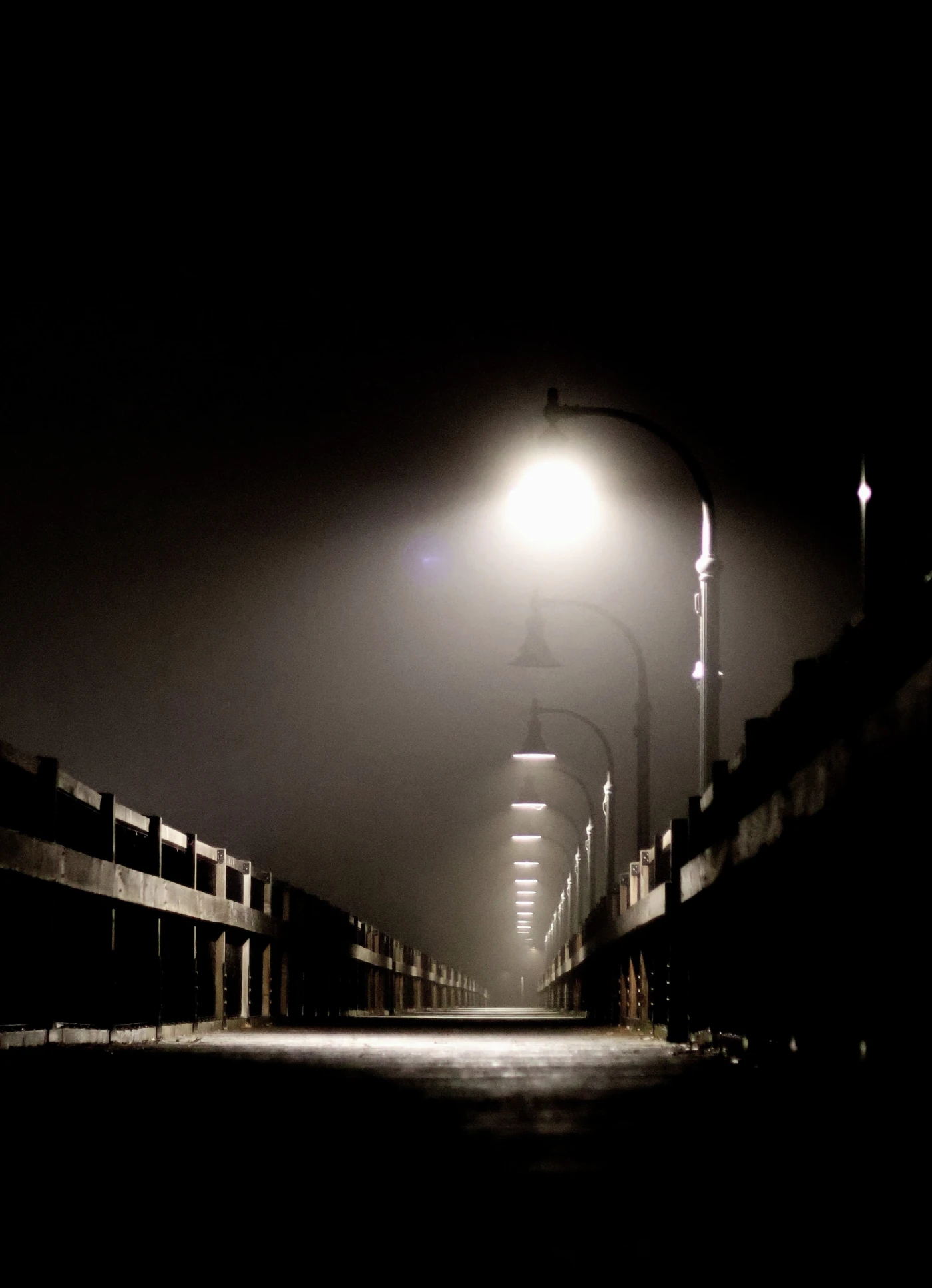 A wooden bridge is lit by a series of lamps on a damp, foggy night. The lights create a repeated pattern down the bridge and into the distance.